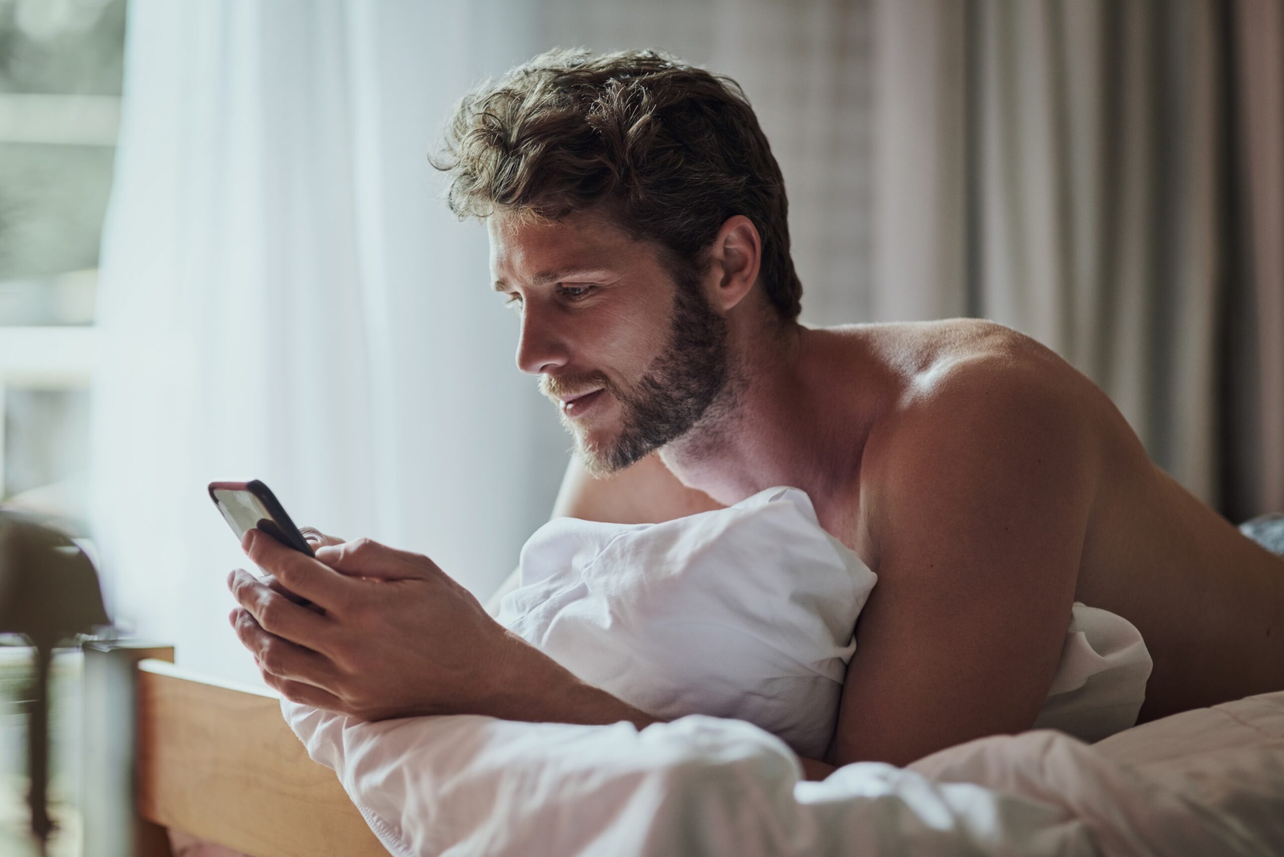 Gay man browsing dating apps while partner sleeps, illustrating digital temptations that lead to infidelity requiring couples counseling.